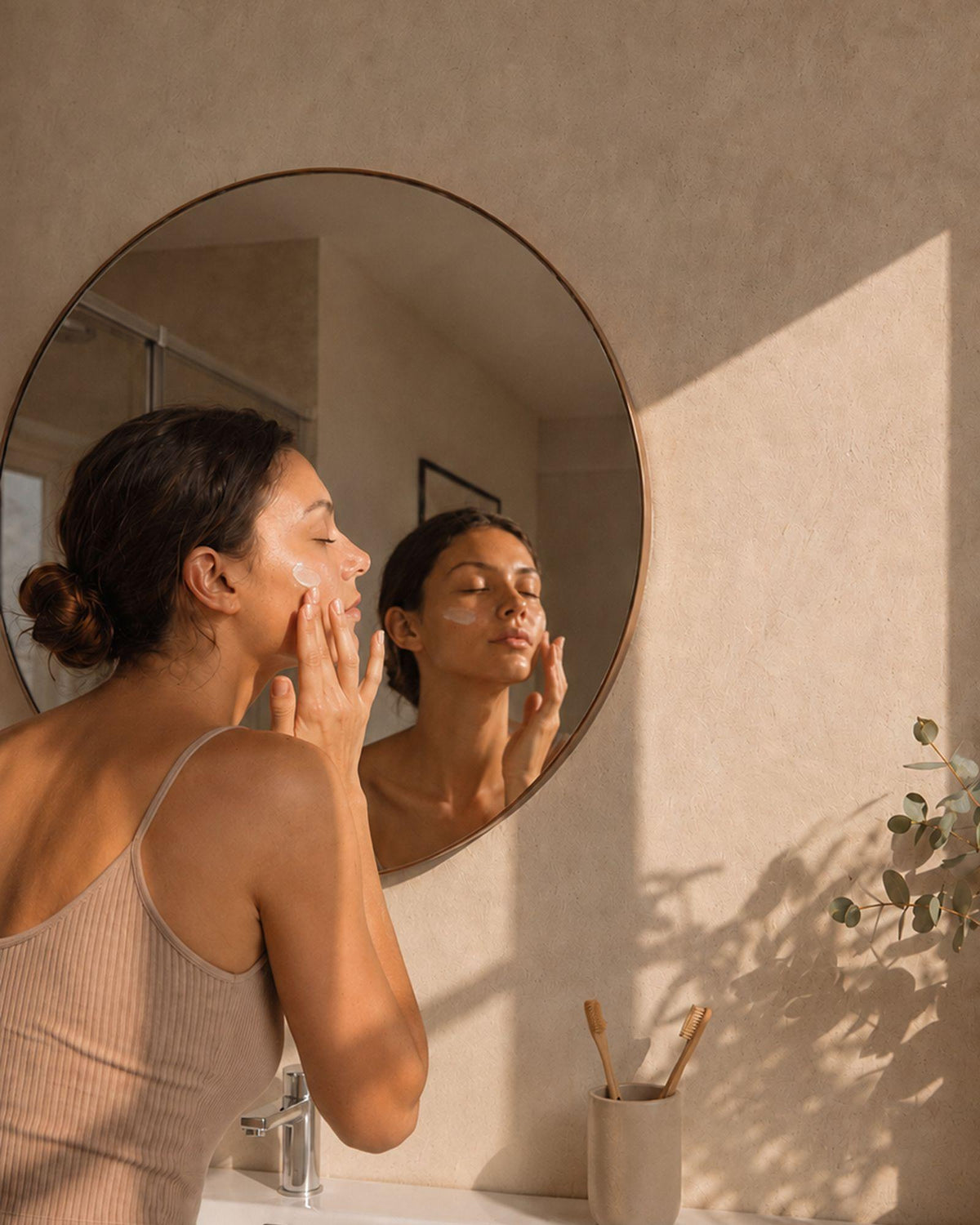 Femme appliquant un soin du visage devant son miroir dans une salle de bain lumineuse.